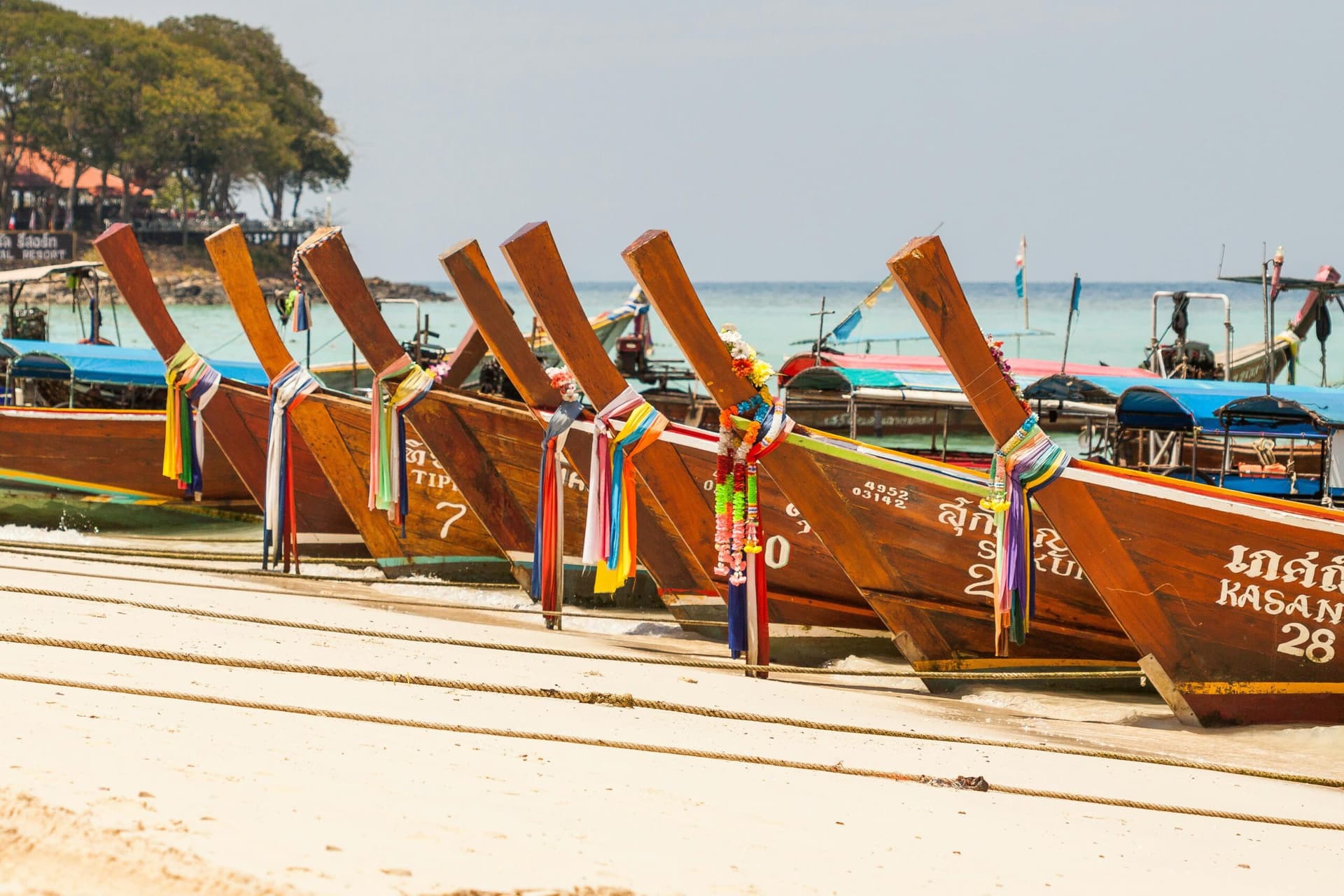 Row of colorful Thai long-tail boats decorated with ribbons and garlands on a sandy beach with turquoise water in the background